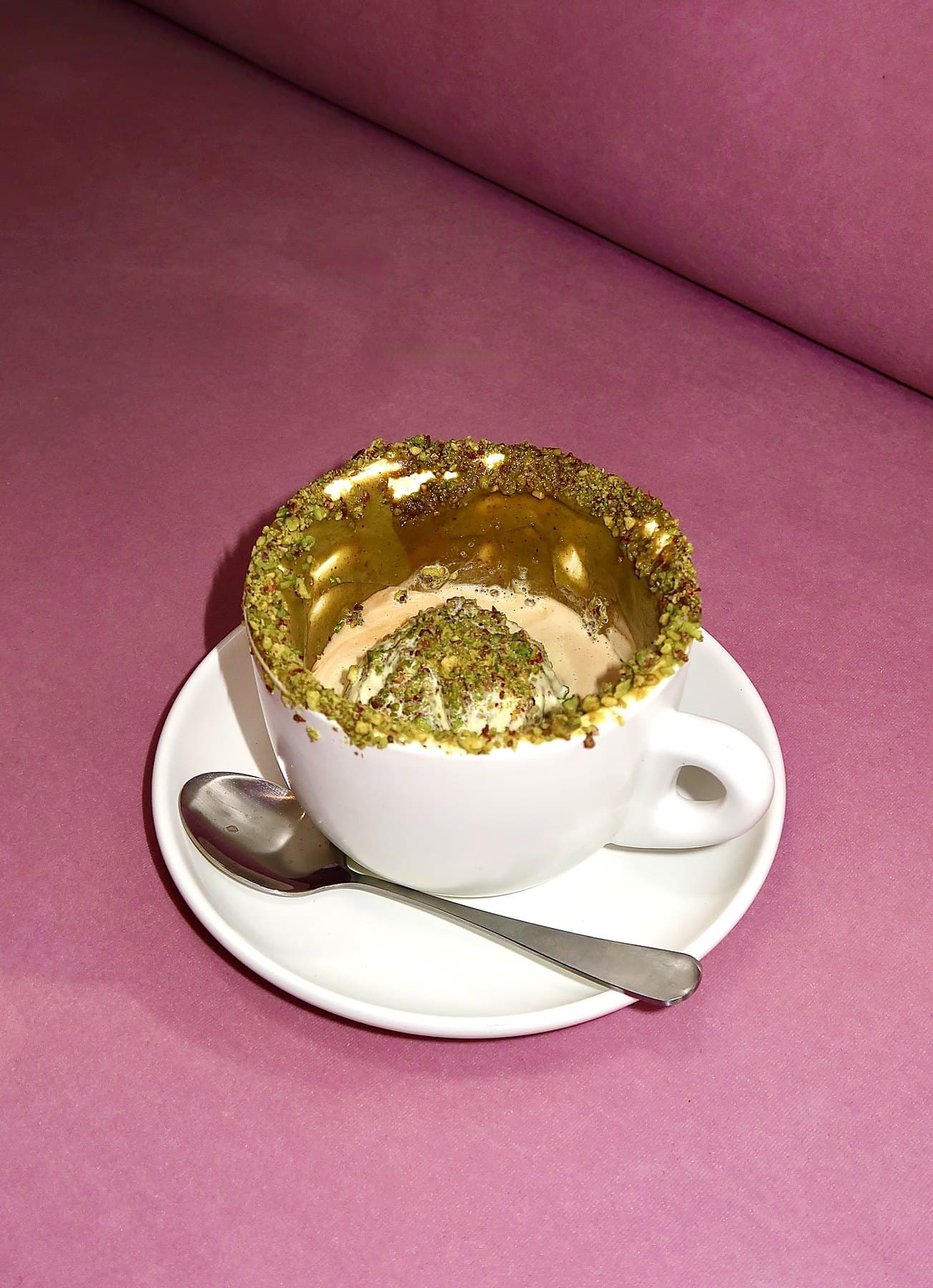 Pistachio affogato in a white cup with a crushed pistachio rim on a pink velvet seat